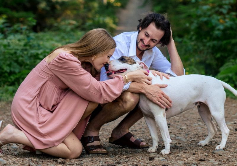 Woman and man kneel near their dog outdoors at a forest trail